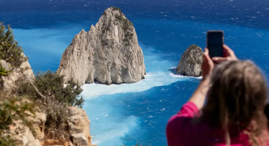 Widok na turkusowe morze i skaliste wysepki. Kobieta robi zdjęcie telefonem. Zakynthos czy warto? Tak, warto!