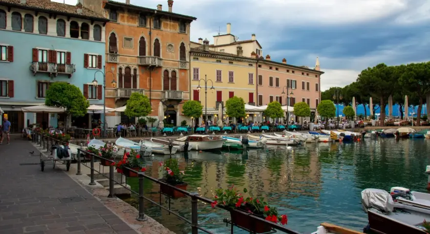 Kolorowe kamienice nad jeziorem w Desenzano del Garda, z łodziami zacumowanymi przy brzegu i stolikami restauracyjnymi na promenadzie.