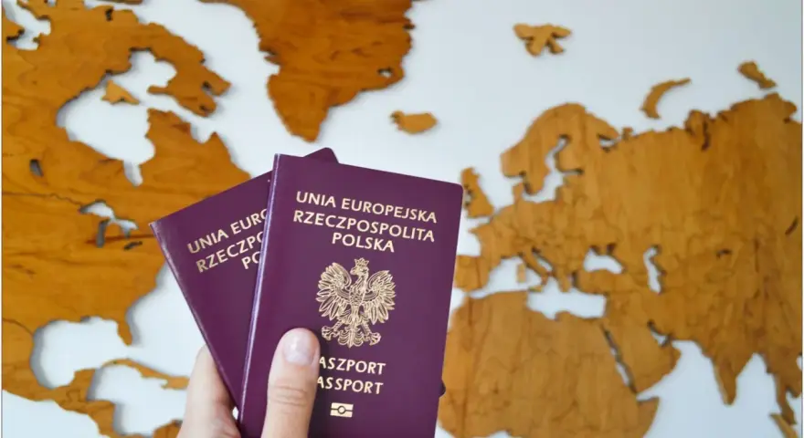 Dwa polskie paszporty w dłoni na tle drewnianej mapy świata. Planujesz podróż? Dowiedz się, jak wyrobić paszport.