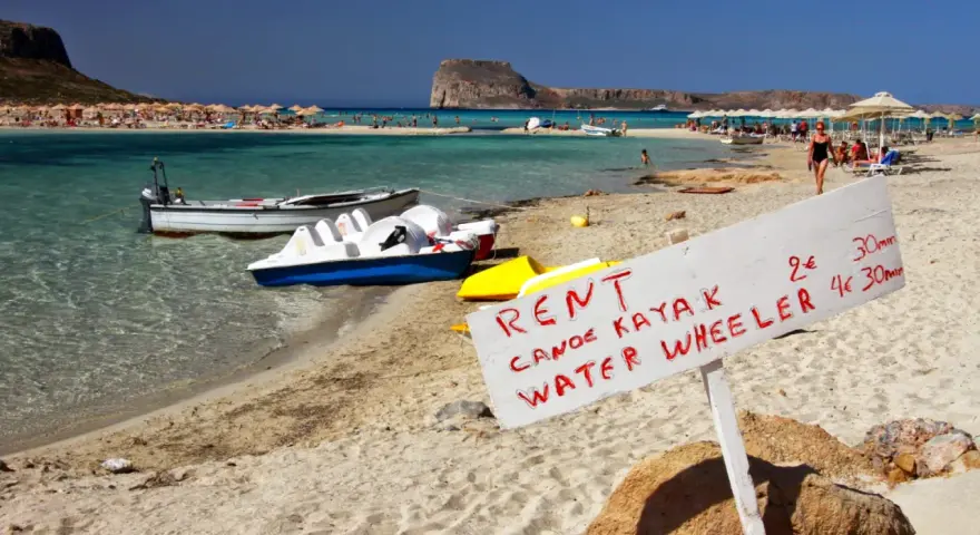 Maj na Krecie: plaża z leżakami, turkusowa woda, łódki i tablica z ofertą wynajmu kajaków i rowerów wodnych.