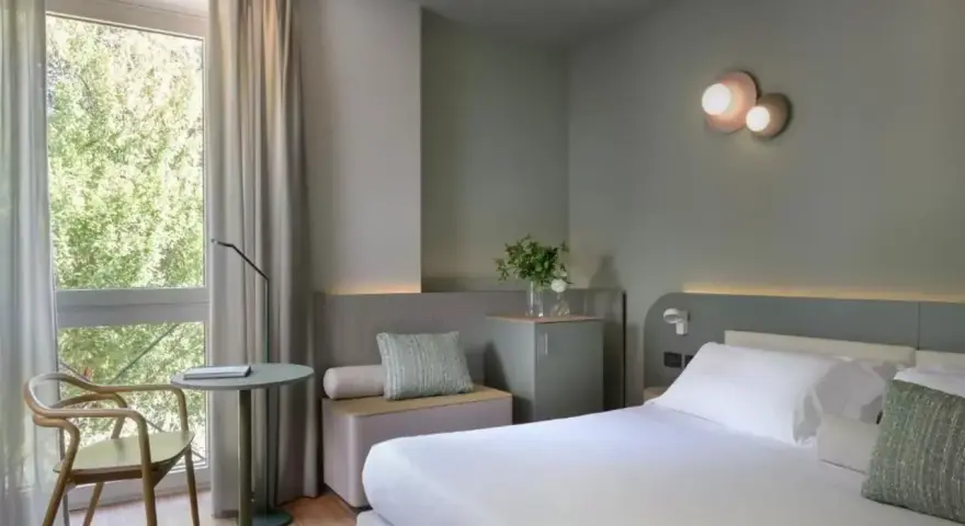 Nowoczesny pokój hotelowy z łóżkiem, fotelem i oknem z widokiem na zieleń.