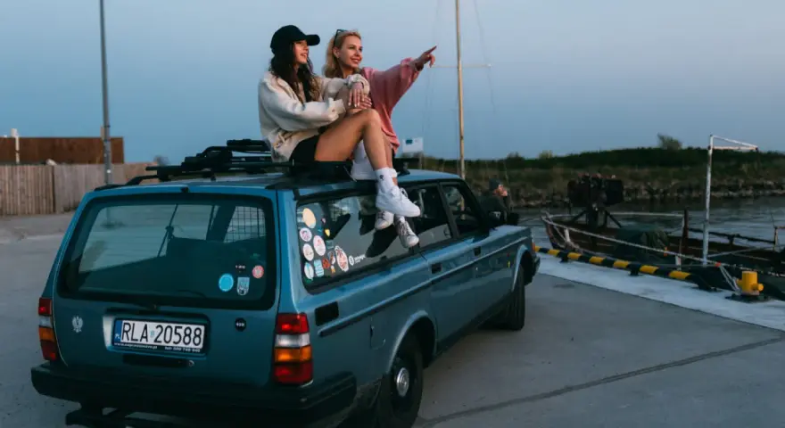 Dwie kobiety siedzą na dachu niebieskiego kombi przy nabrzeżu, jedna wskazuje palcem w dal.