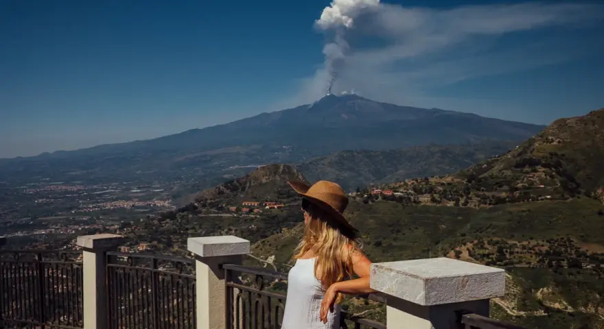 Kobieta w kapeluszu patrzy na dymiący wulkan Etna z tarasu widokowego.