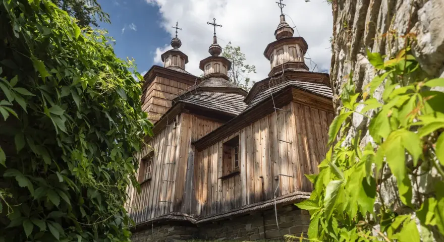 Drewniana cerkiew z trzema kopułami, otoczona zielenią, na tle błękitnego nieba z chmurami.