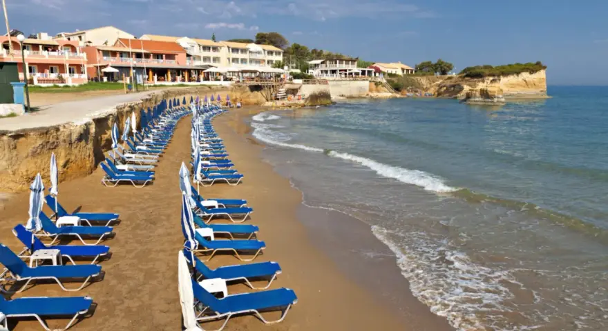 Rzędy leżaków i parasoli na piaszczystej plaży, z hotelami w tle. Idealne miejsce, by odpocząć i zastanowić się, gdzie na Korfu wybrać się na wakacje.