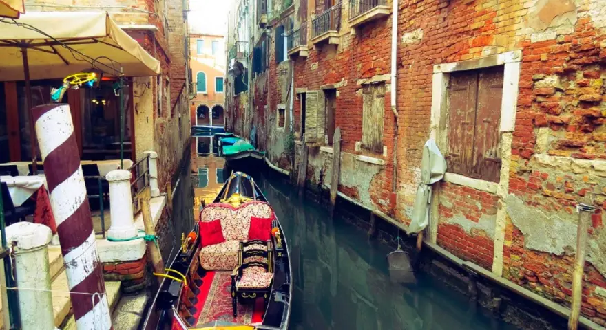 Gondola z czerwonymi poduszkami i dywanem w wąskim kanale Wenecji, otoczonym starymi ceglanymi budynkami.