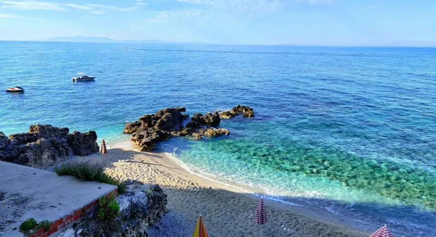 Albania, czerwiec: słoneczna pogoda nad krystalicznie czystym morzem, kamienista plaża z parasolami i łódkami na horyzoncie.