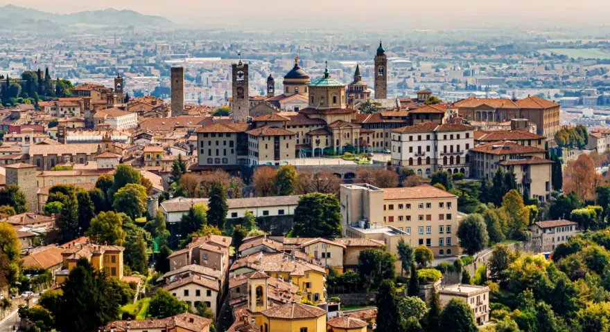 Widok na **bergamo stare miasto** z wieżami i kopułami, otoczone zielenią i innymi budynkami.