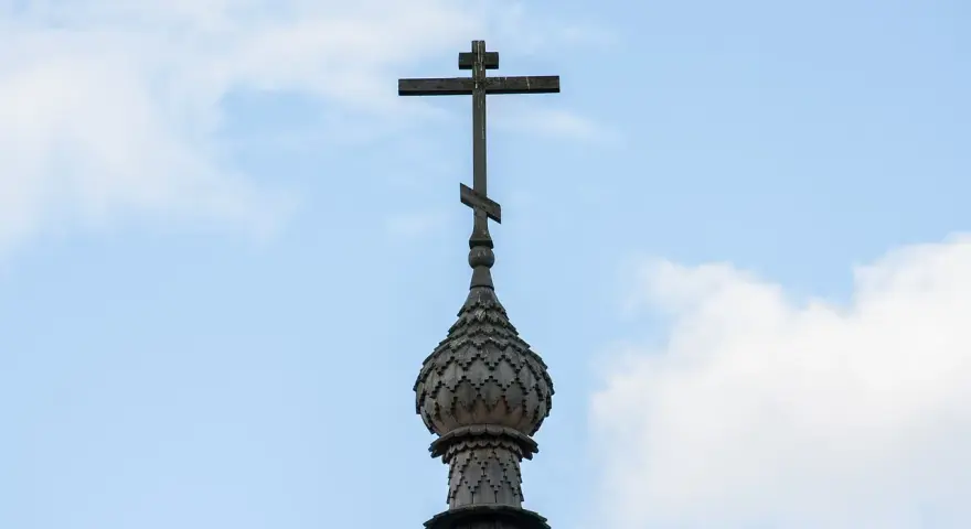 Drewniany krzyż prawosławny na tle błękitnego nieba z chmurami.
