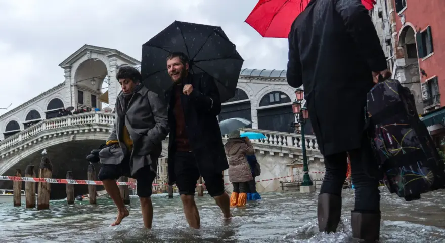 Ludzie brodzą w wodzie podczas acqua alta w Wenecji. Most Rialto w tle, deszcz i parasole.