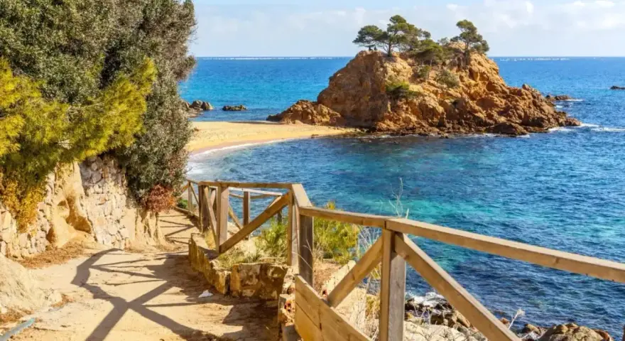 Ścieżka z drewnianą balustradą prowadzi do urokliwej zatoczki z piaszczystą plażą i skalistą wysepką. To idealne miejsce na odkrywanie uroków Costa Brava.