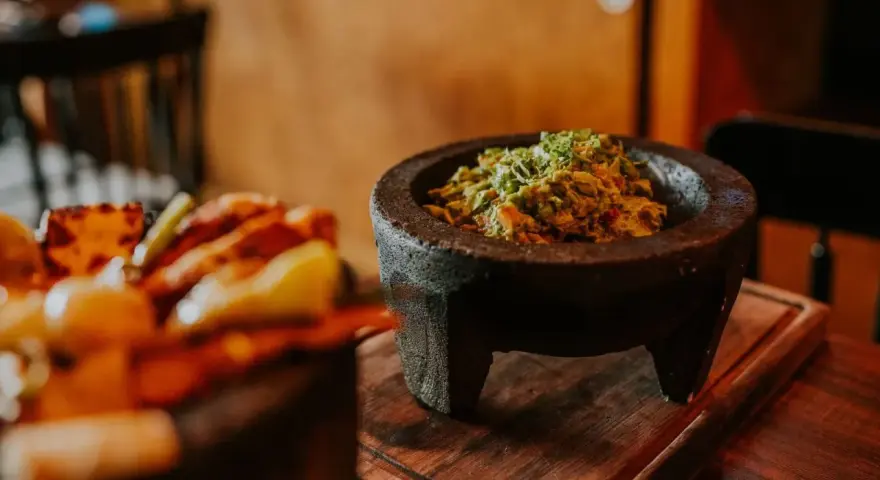 Świeże guacamole w kamiennym moździerzu, obok talerza z innymi najpopularniejszymi daniami meksykańskimi.