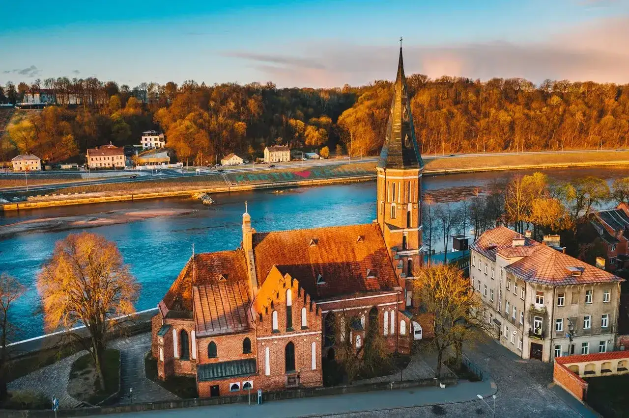 Gotycka ceglana katedra nad rzeką, otoczona jesiennymi drzewami i zabudowaniami. To widok, z czego słynie Litwa - piękno architektury i natury.