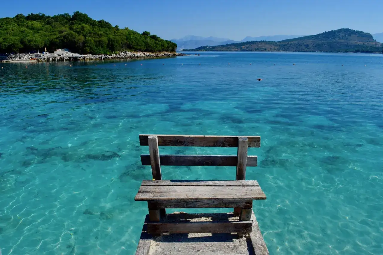 Albania, lipiec. Słoneczna pogoda nad krystalicznie czystą wodą. Pusta ławka zaprasza do podziwiania widoku.