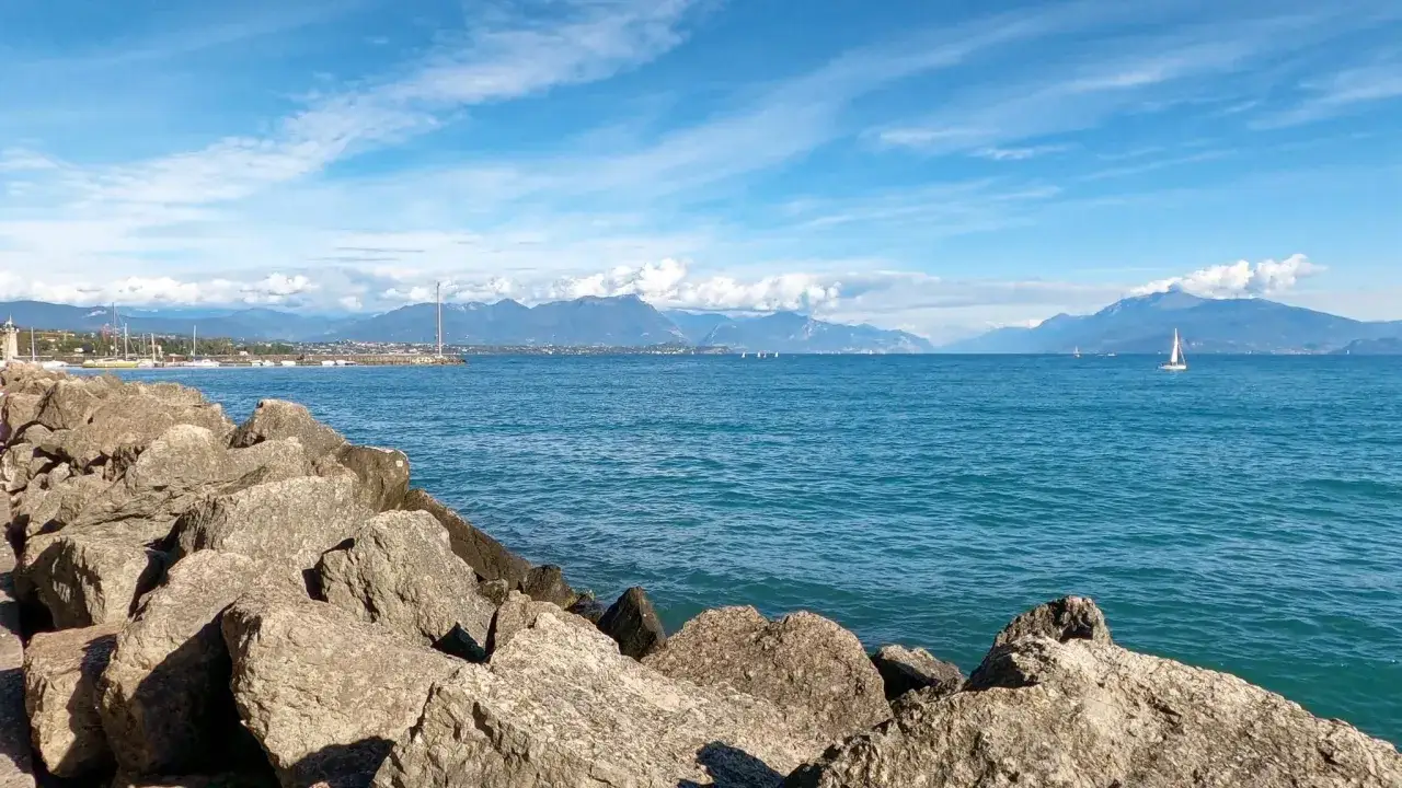 Kamieniste wybrzeże w Desenzano del Garda, błękitne jezioro, góry w oddali i żaglówki na wodzie.