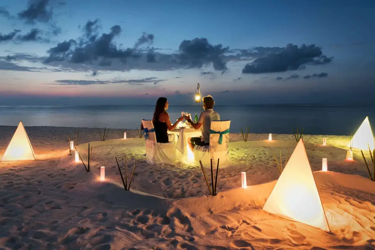Romantyczna kolacja na plaży, idealne miejsce na podr&oacute;ż poślubną. Para wznosi toasty przy blasku lampion&oacute;w.