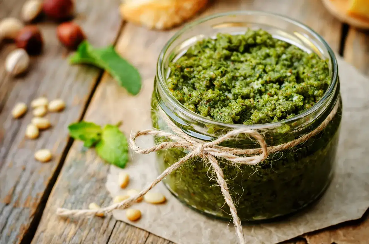 Słoik domowego pesto bazyliowego, ozdobiony sznurkiem. Obok orzeszki piniowe i listki bazylii. Idealny przepis na pesto bazyliowe.