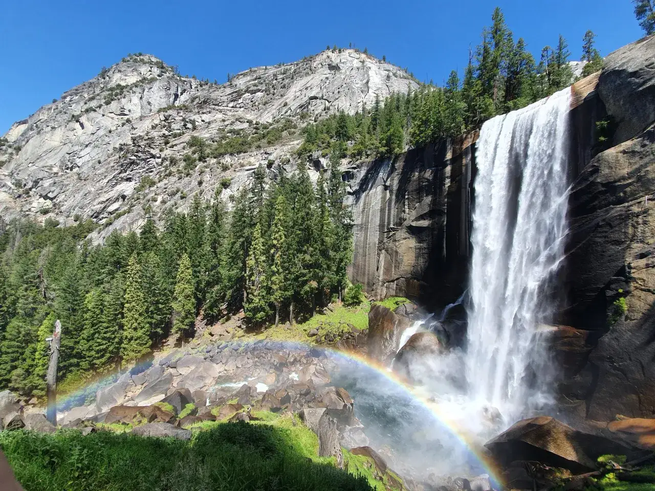 Wodospad Vernal Falls w parku narodowym Yosemite, z tęczą tworzącą się w rozpryskach wody. Majestatyczna skała i zielone drzewa w tle.
