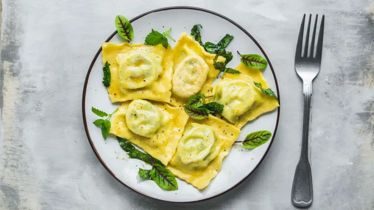 Pyszne ravioli z ricottą i szpinakiem, polane sosem i udekorowane świeżymi ziołami. Obok leży widelec.