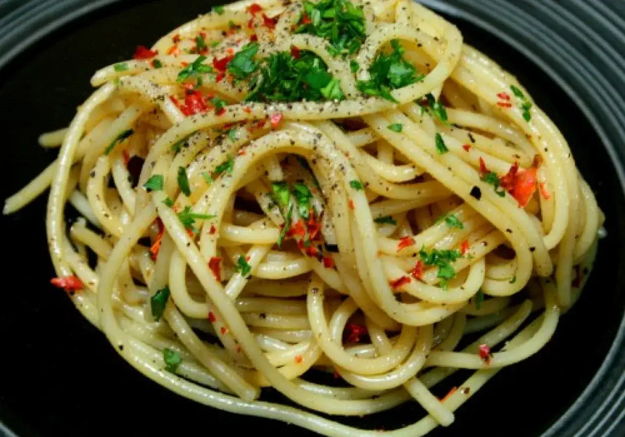 Pyszny makaron aglio olio, posypany natką pietruszki i płatkami chili, podany na czarnym talerzu.