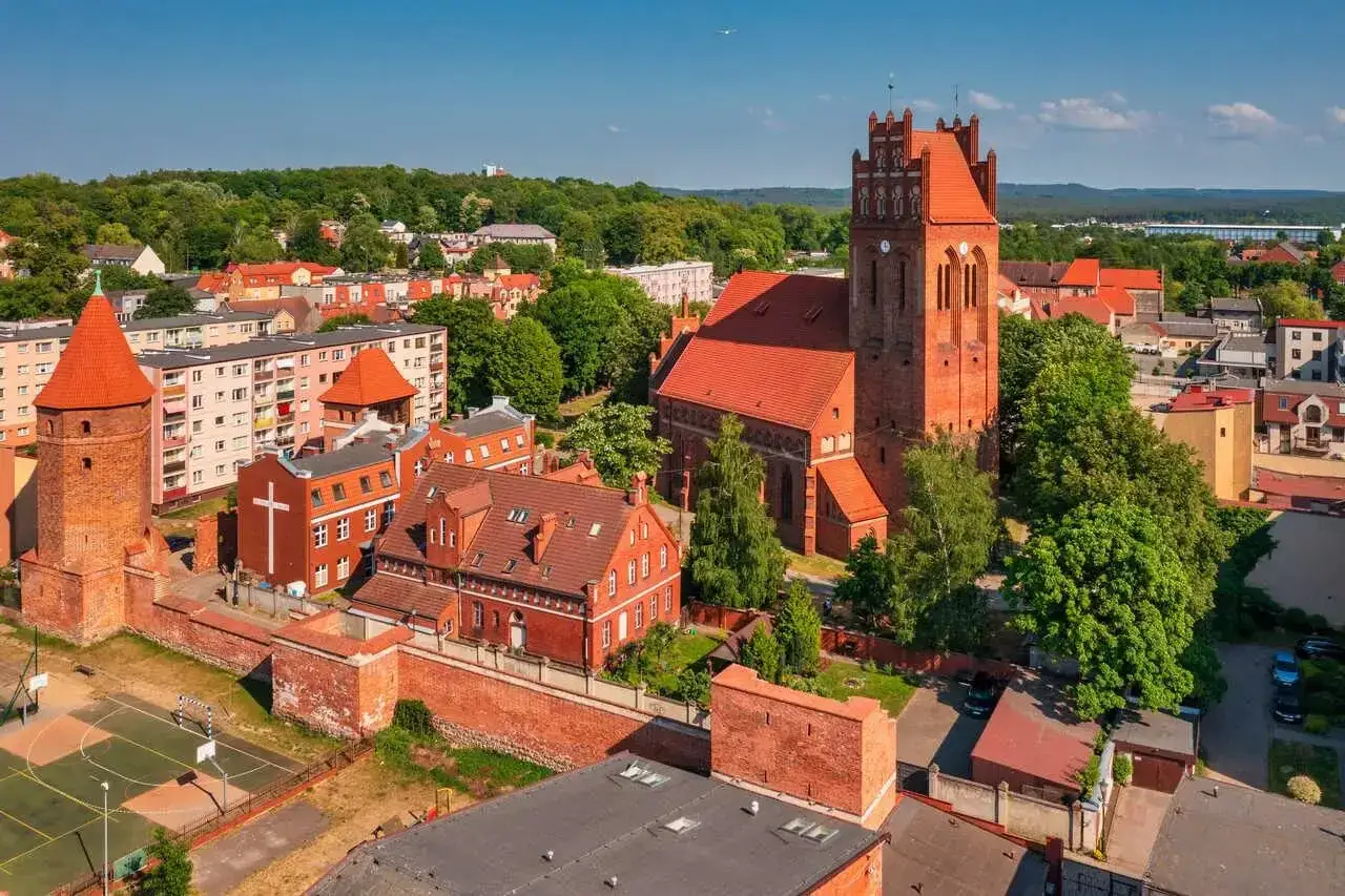 Zamek krzyżacki w Lęborku, z charakterystyczną wieżą i ceglaną architekturą, otoczony zielenią i miejską zabudową.