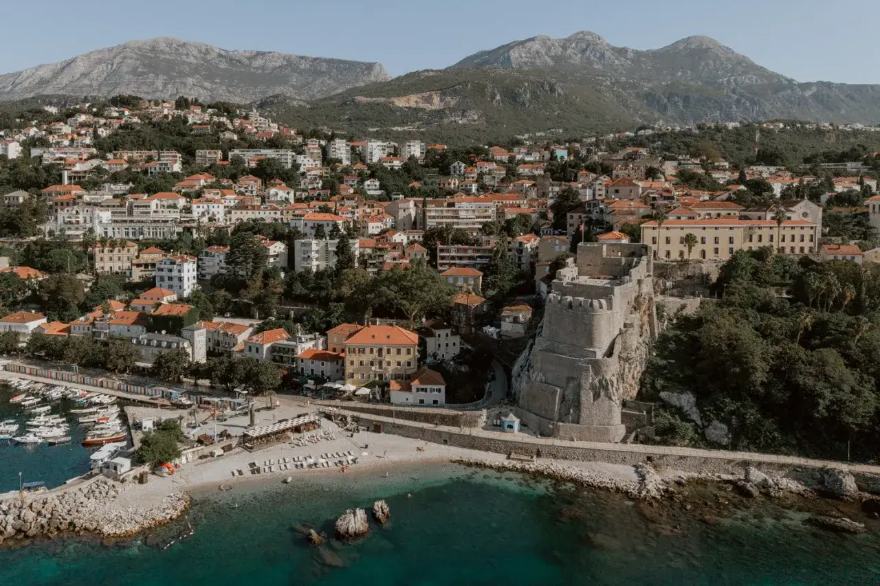 Widok na Herceg Novi plaże i zabytkową twierdzę na klifie. W tle góry i malownicze miasteczko z czerwonymi dachami.