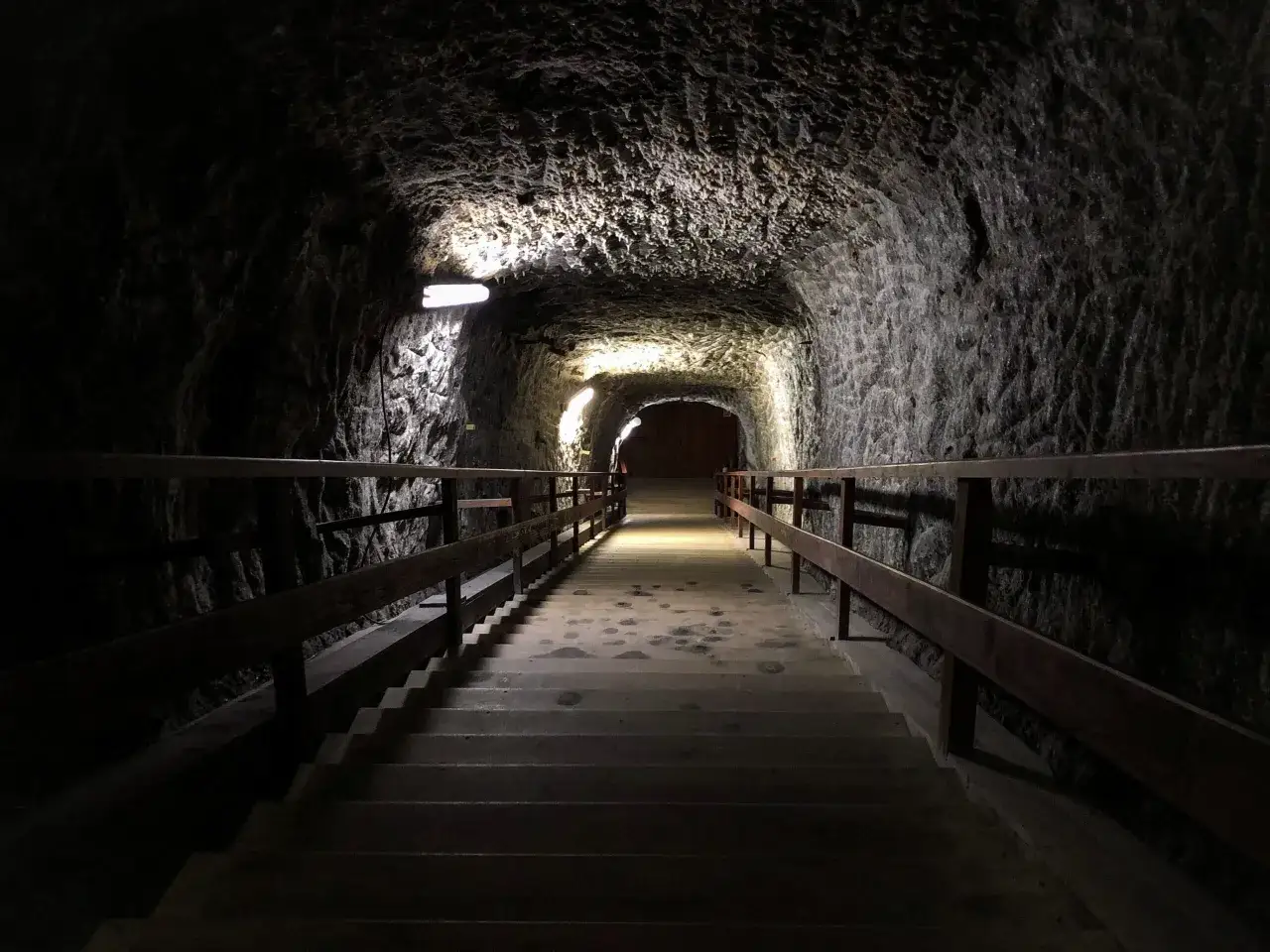 Kamienne schody w kopalni w Wieliczce, gdzie kiedyś wydobywano sól. Mroczny tunel oświetlony lampami, z drewnianymi poręczami.
