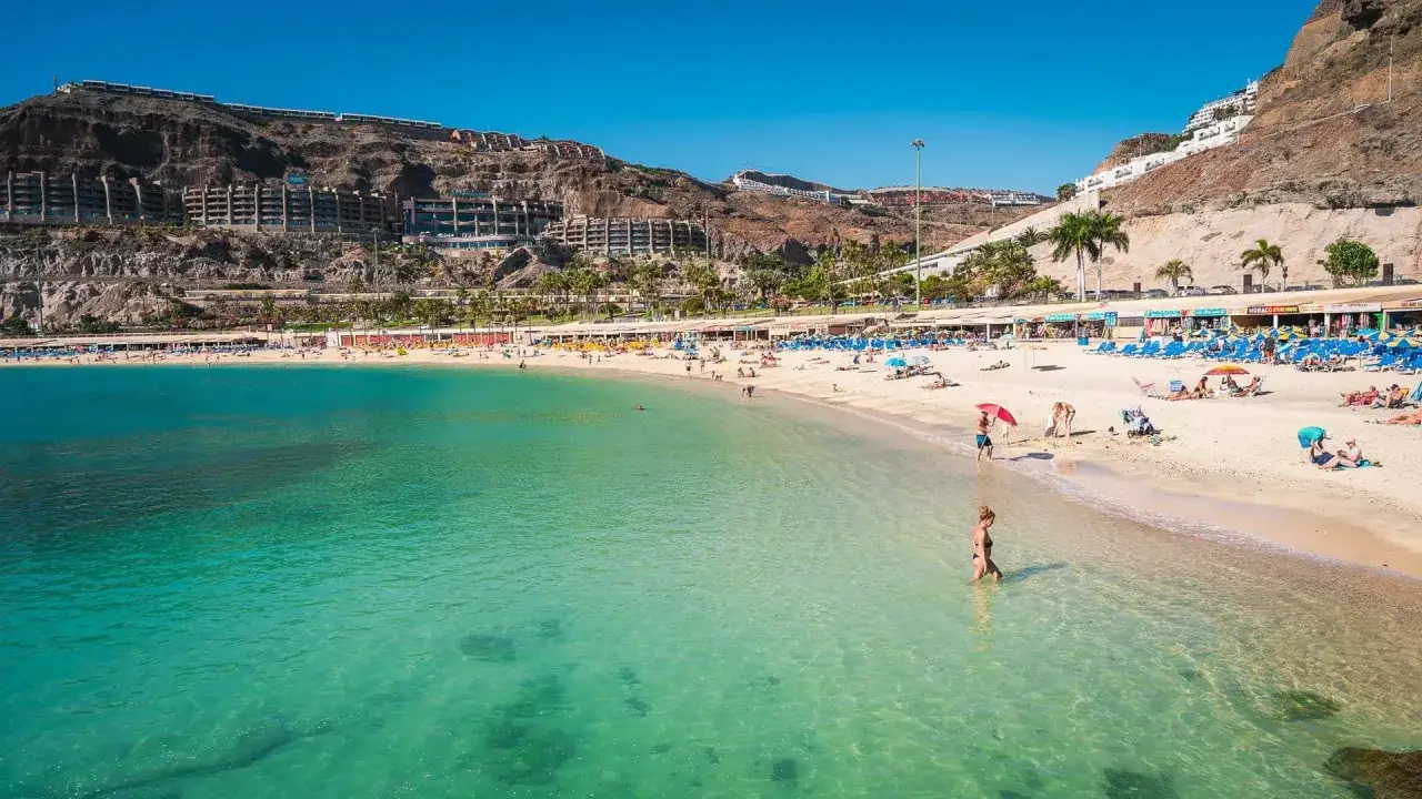 Gran Canaria pogoda w styczniu: słoneczna plaża z turkusową wodą, ludźmi odpoczywającymi na piasku i hotelami na zboczu góry.