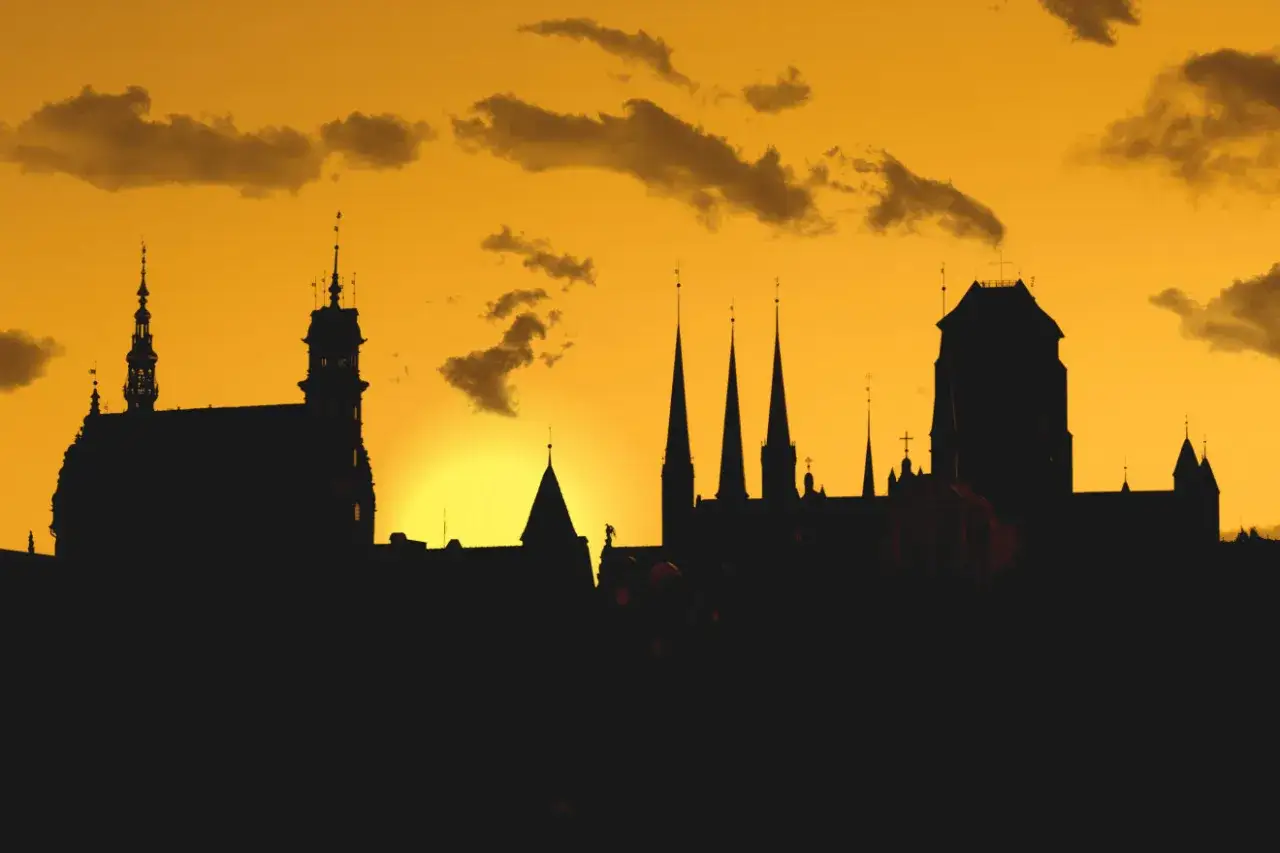 Zachód słońca nad miastem, idealne miejsce gdzie na city break. Sylwetki kościołów i kamienic na tle pomarańczowego nieba.