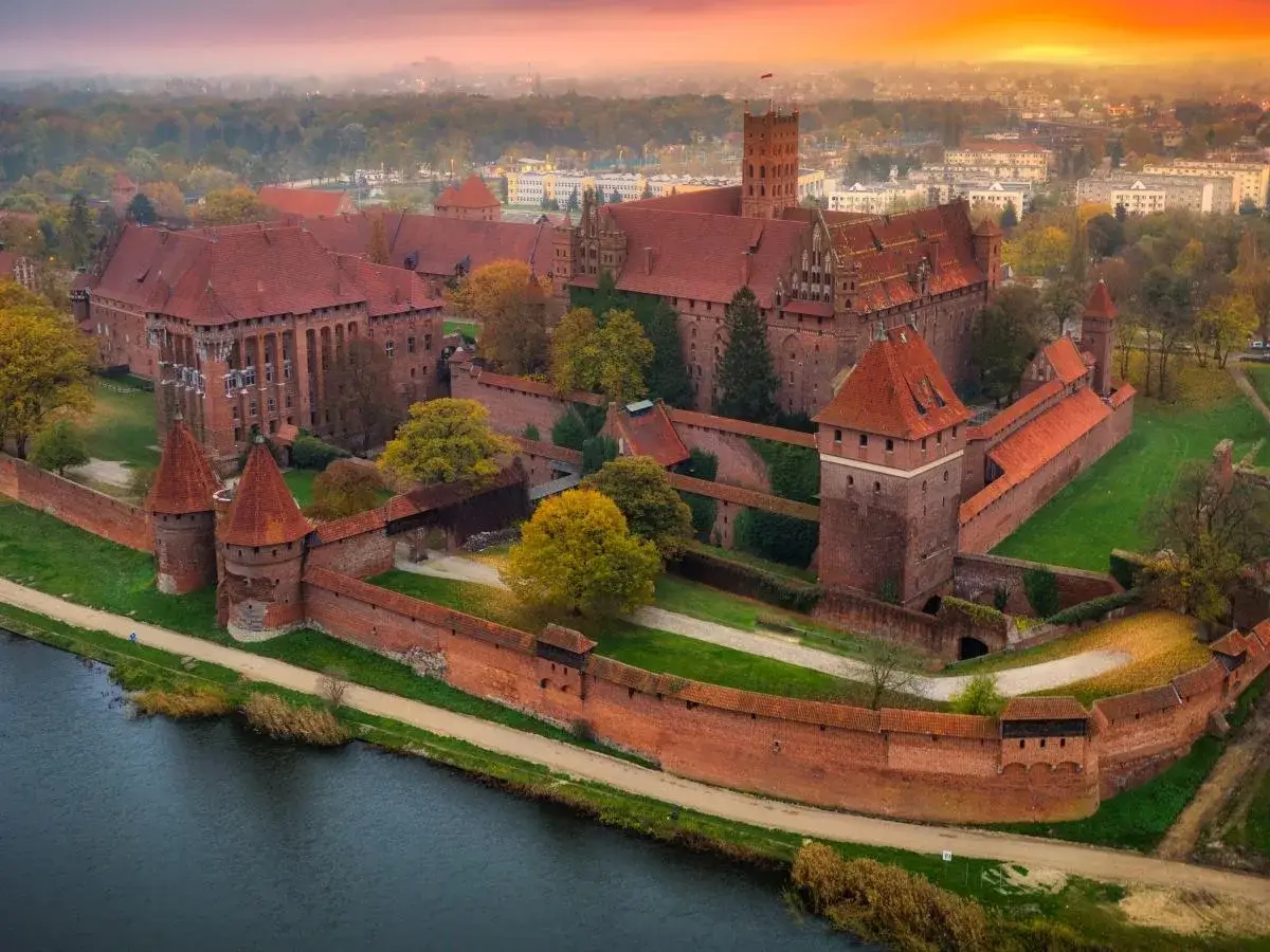 Potężny zamek w Malborku, którego budowa rozpoczęła się w XIII wieku, otoczony ceglanymi murami i wieżami, lśni w blasku zachodzącego słońca.