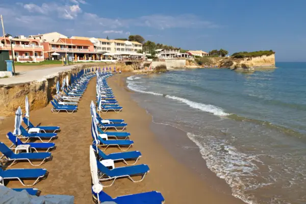Rzędy leżaków i parasoli na piaszczystej plaży, z hotelami w tle. Idealne miejsce, by odpocząć i zastanowić się, gdzie na Korfu wybrać się na wakacje.