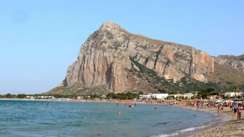 Słoneczna plaża w San Vito Lo Capo, z turkusową wodą i majestatyczną górą w tle. Tłumy ludzi cieszą się wakacjami.