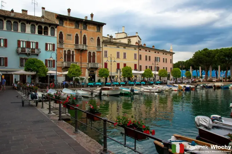 Kolorowe kamienice nad jeziorem w Desenzano del Garda, z łodziami zacumowanymi przy brzegu i stolikami restauracyjnymi na promenadzie.