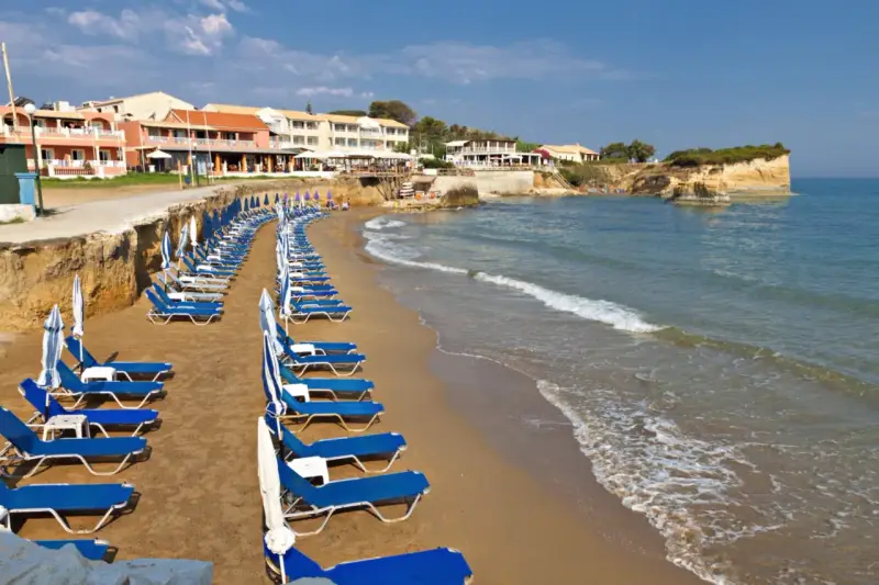 Rzędy leżaków i parasoli na piaszczystej plaży, z hotelami w tle. Idealne miejsce, by odpocząć i zastanowić się, gdzie na Korfu wybrać się na wakacje.