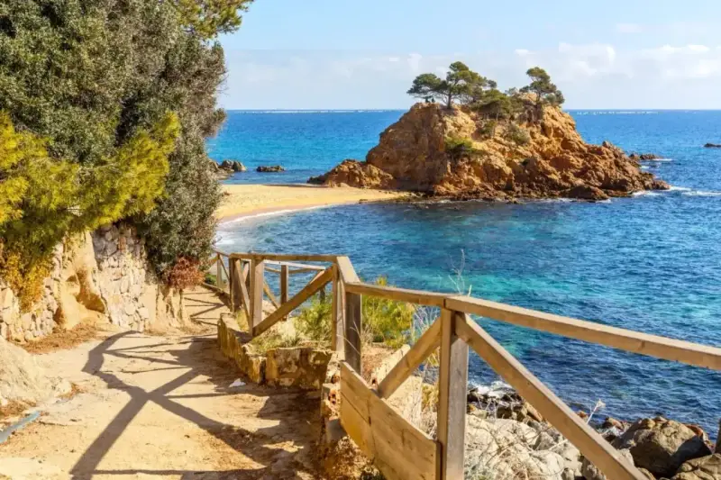 Ścieżka z drewnianą balustradą prowadzi do urokliwej zatoczki z piaszczystą plażą i skalistą wysepką. To idealne miejsce na odkrywanie uroków Costa Brava.